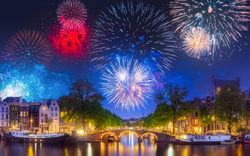 Feuerwerk über einer beleuchteten Stadtlandschaft mit Brücke und Kanal bei Nacht.
