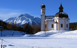 Kirche in verschneiter Alpenlandschaft mit Bergen im Hintergrund.