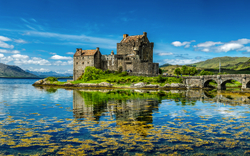 Burg auf Insel mit Brücke und reflektierendem See im Vordergrund.